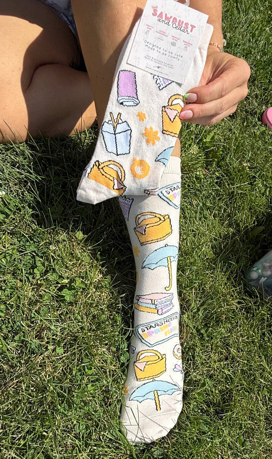 A person sitting on grass holds up a white sock with a colorful illustrated pattern of shopping bags, umbrellas, and gift boxes. The sock has a paper tag attached at the top.