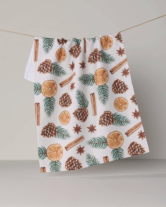 A white kitchen towel with a festive holiday print of pinecones, cinnamon sticks, star anise, evergreen branches, and orange slices, hanging from a string against a neutral gray background.