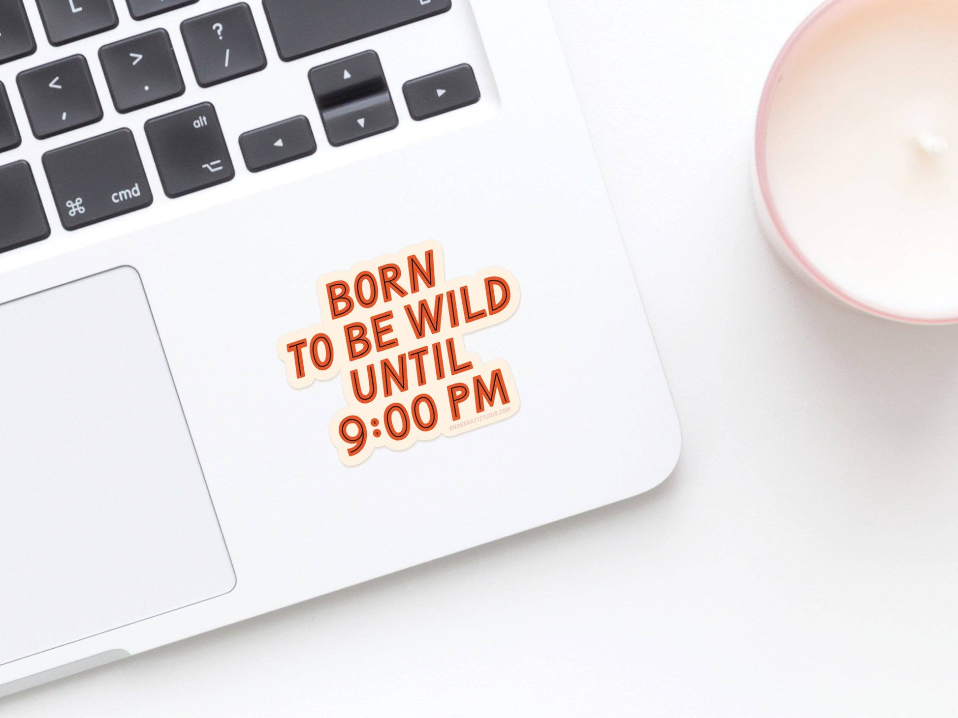 Laptop with 'Born to be wild until 9:00 PM' text next to a candle on a white surface