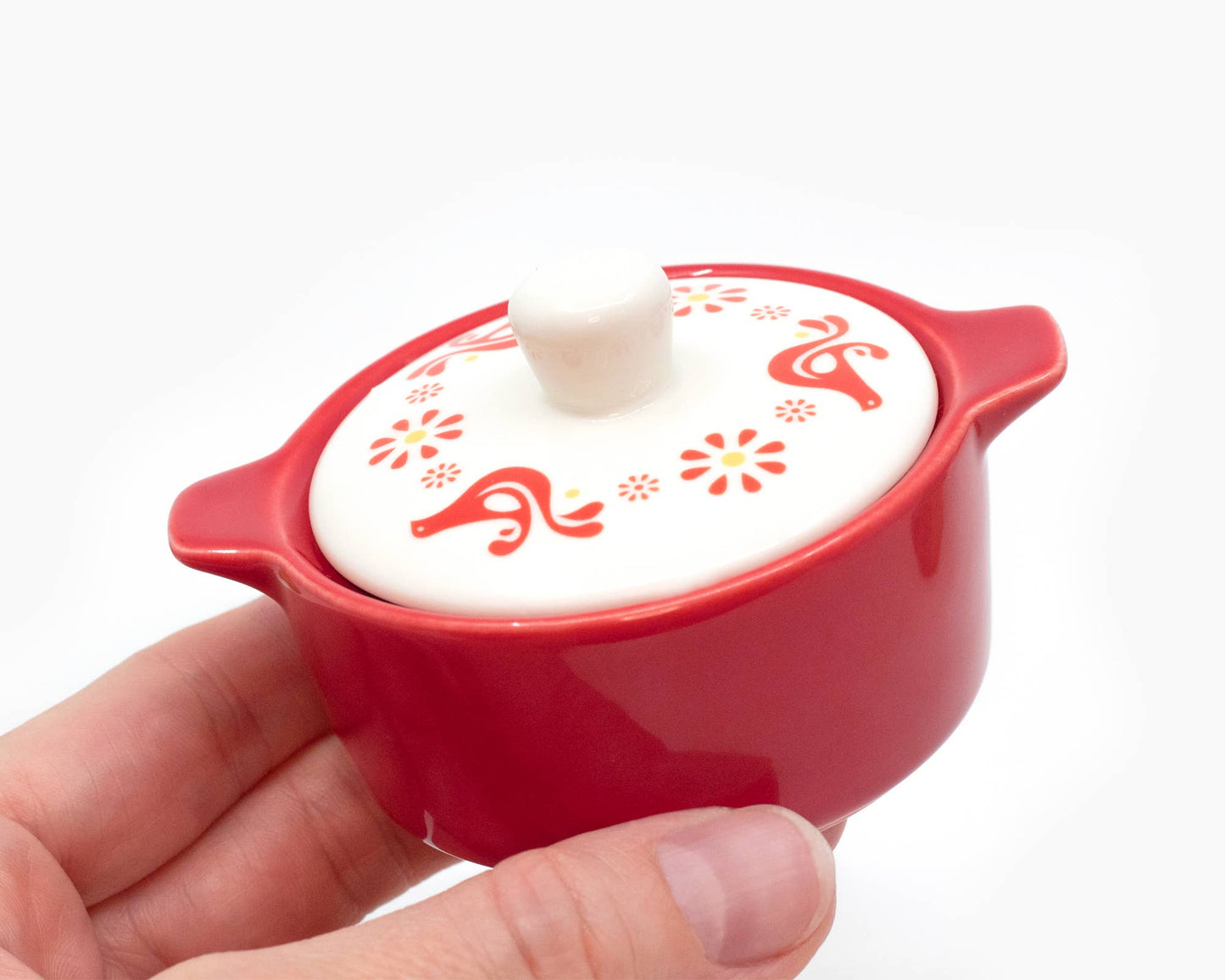Red ceramic pot with white lid held by a hand on a white background