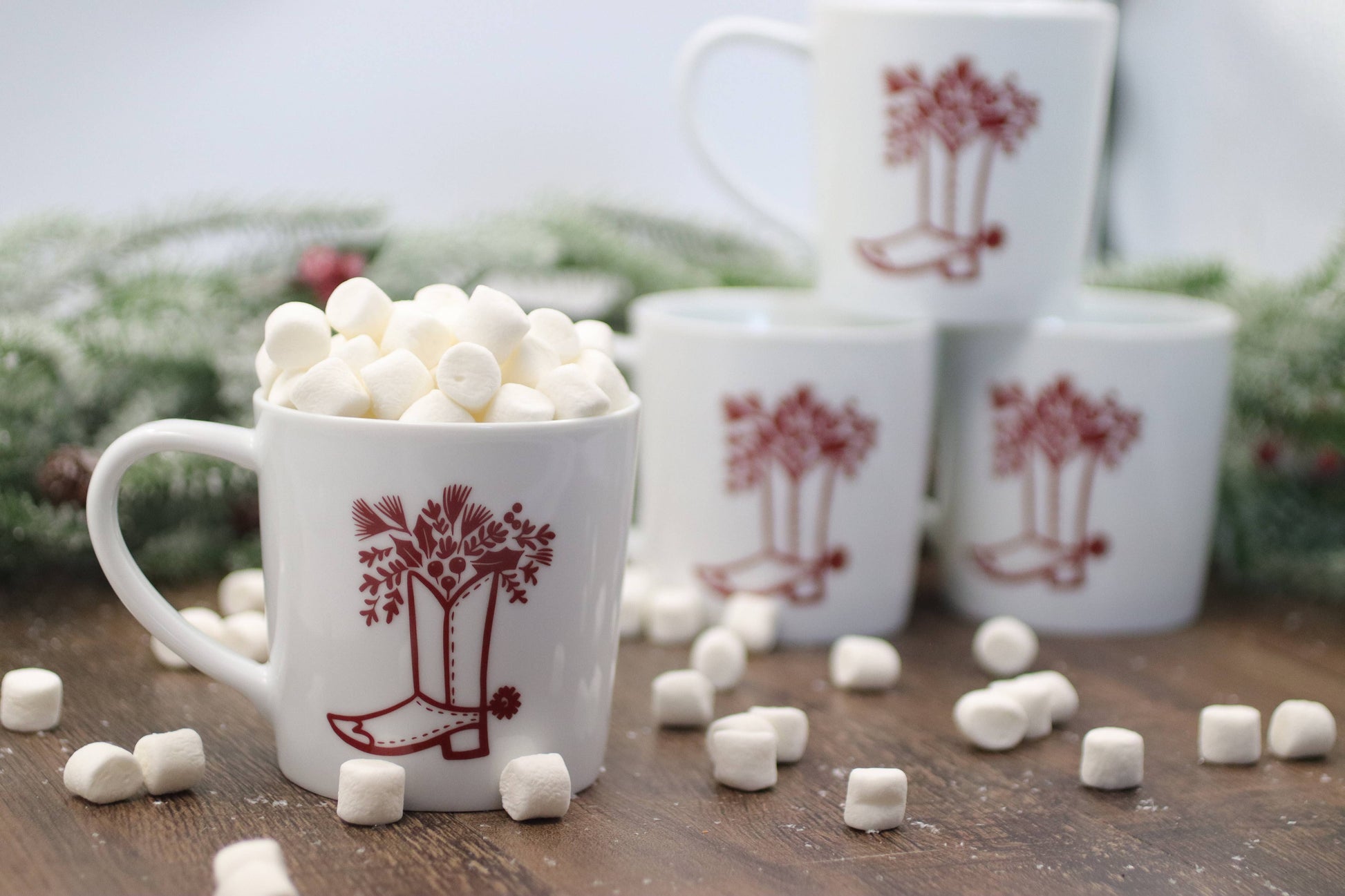 White mugs with red tree design filled with marshmallows on a wooden surface with Christmas decorations.