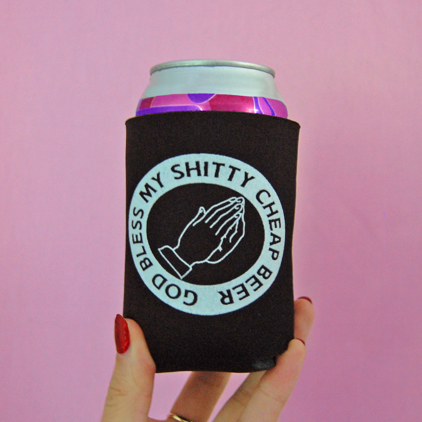 Black can cozy with a design of two praying hands and text saying "God Bless My Shitty Cheap Beer"  held against a pink background