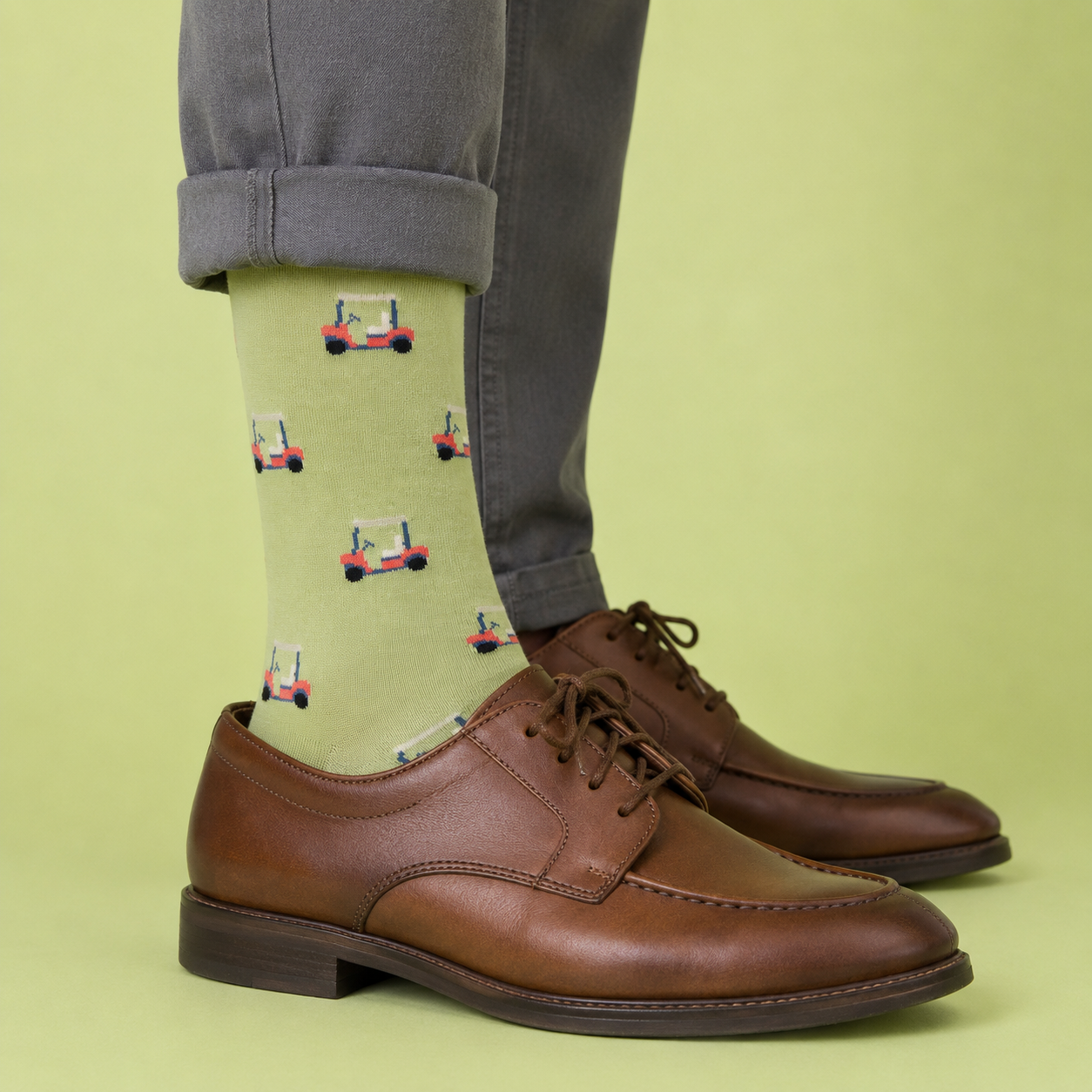 A person wearing light green crew socks with a repeating pattern of red and blue golf carts, paired with brown leather oxford shoes and gray cuffed trousers, set against a solid light green background.