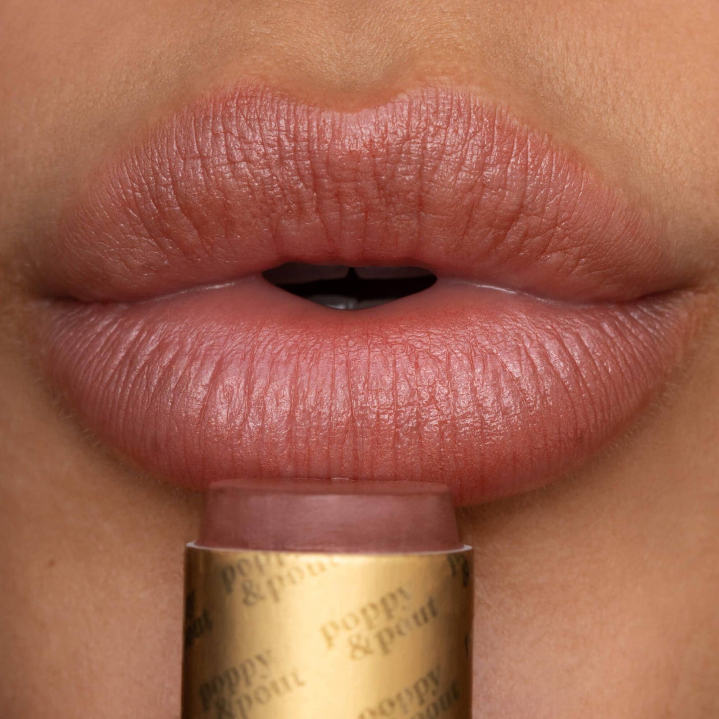 Close-up of a person applying pink lipstick to their lips with a gold tube labeled 'Poppy & Plum'.