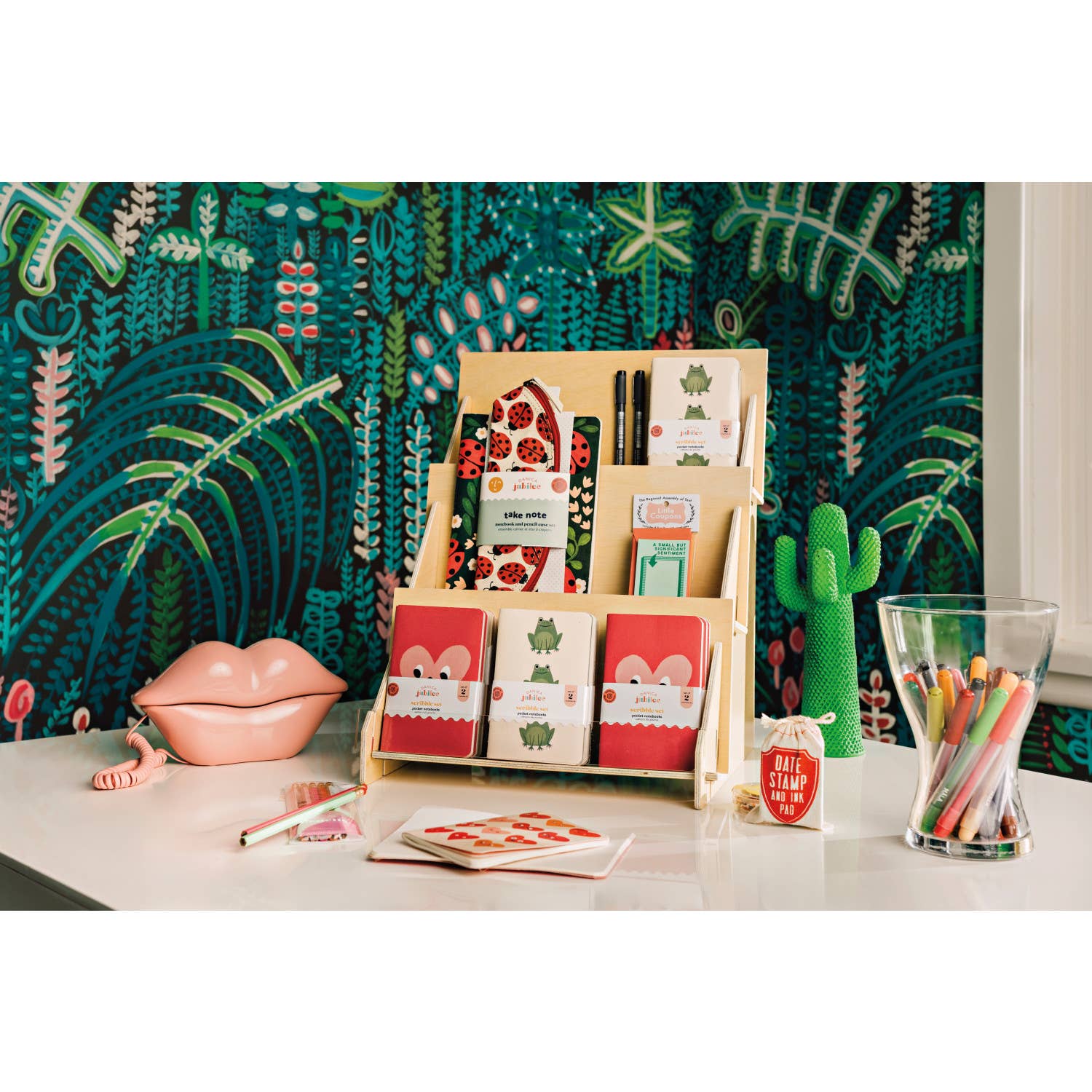 Desk with stationery items including notebooks, pens, and a glass container against a colorful wall.
