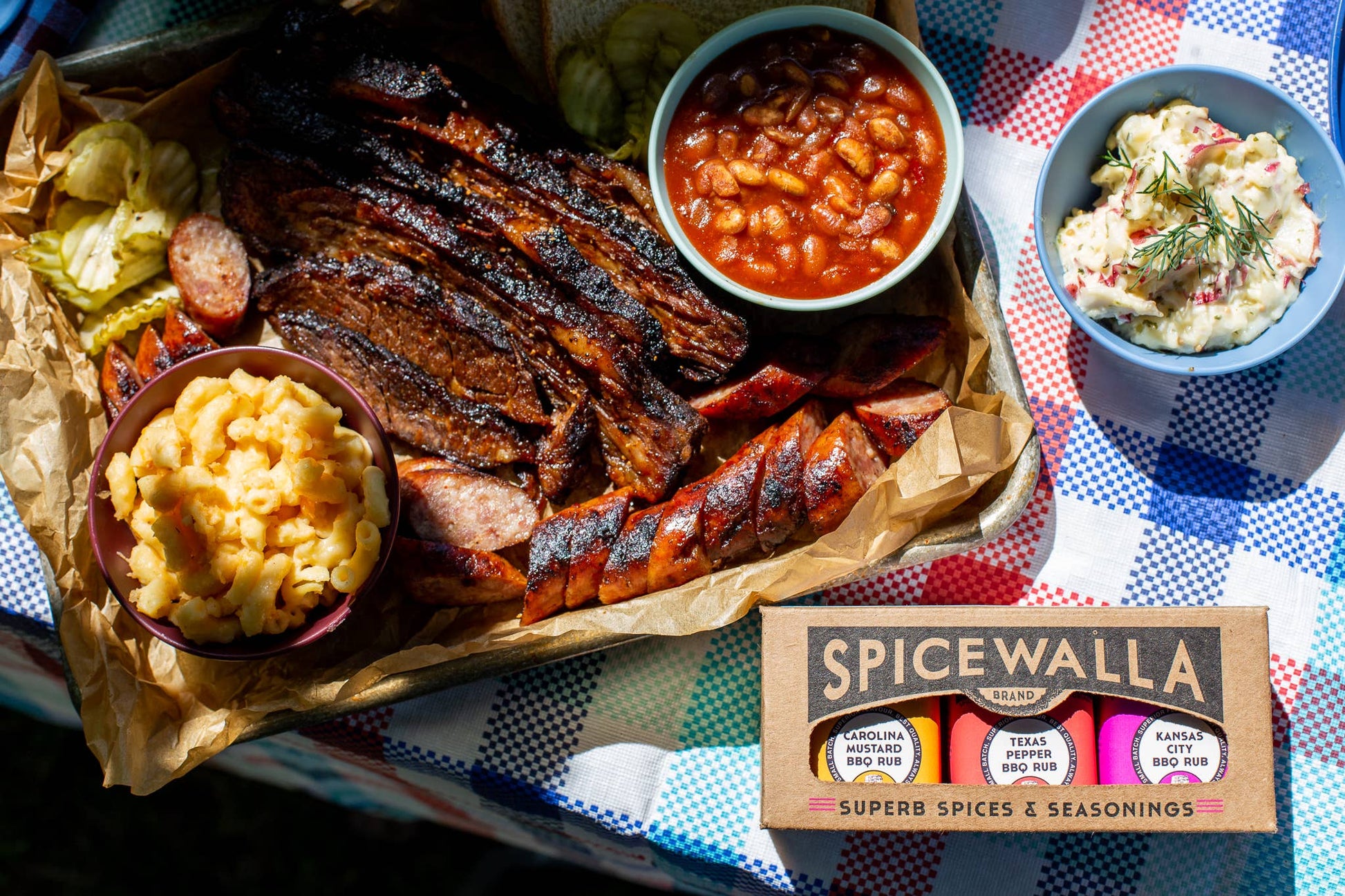 Platter of barbecue meats with sides and Spicewalla spice set on a checkered tablecloth.