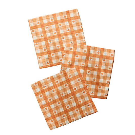 Three square cotton napkins with an orange and white gingham check pattern and small floral motifs, arranged on a light wood surface.