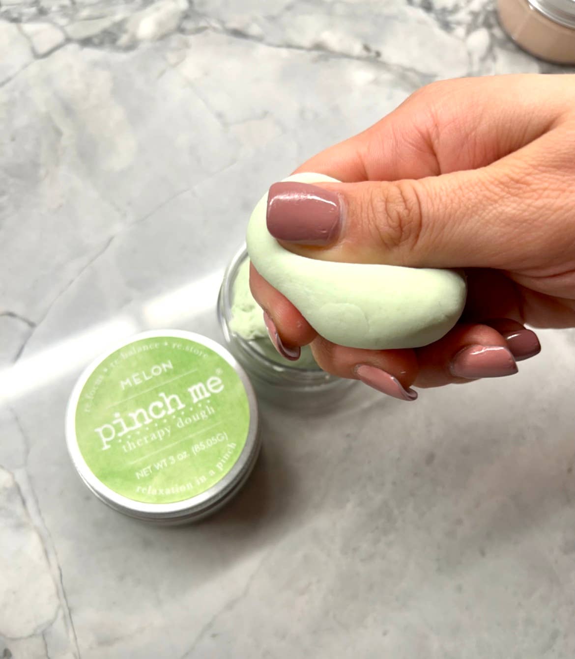 Hand holding a green dough with 'pinch me' container on a marble surface