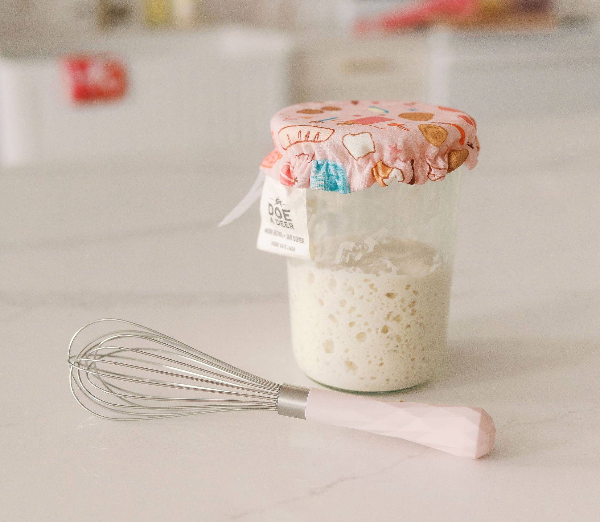 Jar of cottage cheese with a floral lid on a kitchen counter, accompanied by a whisk.