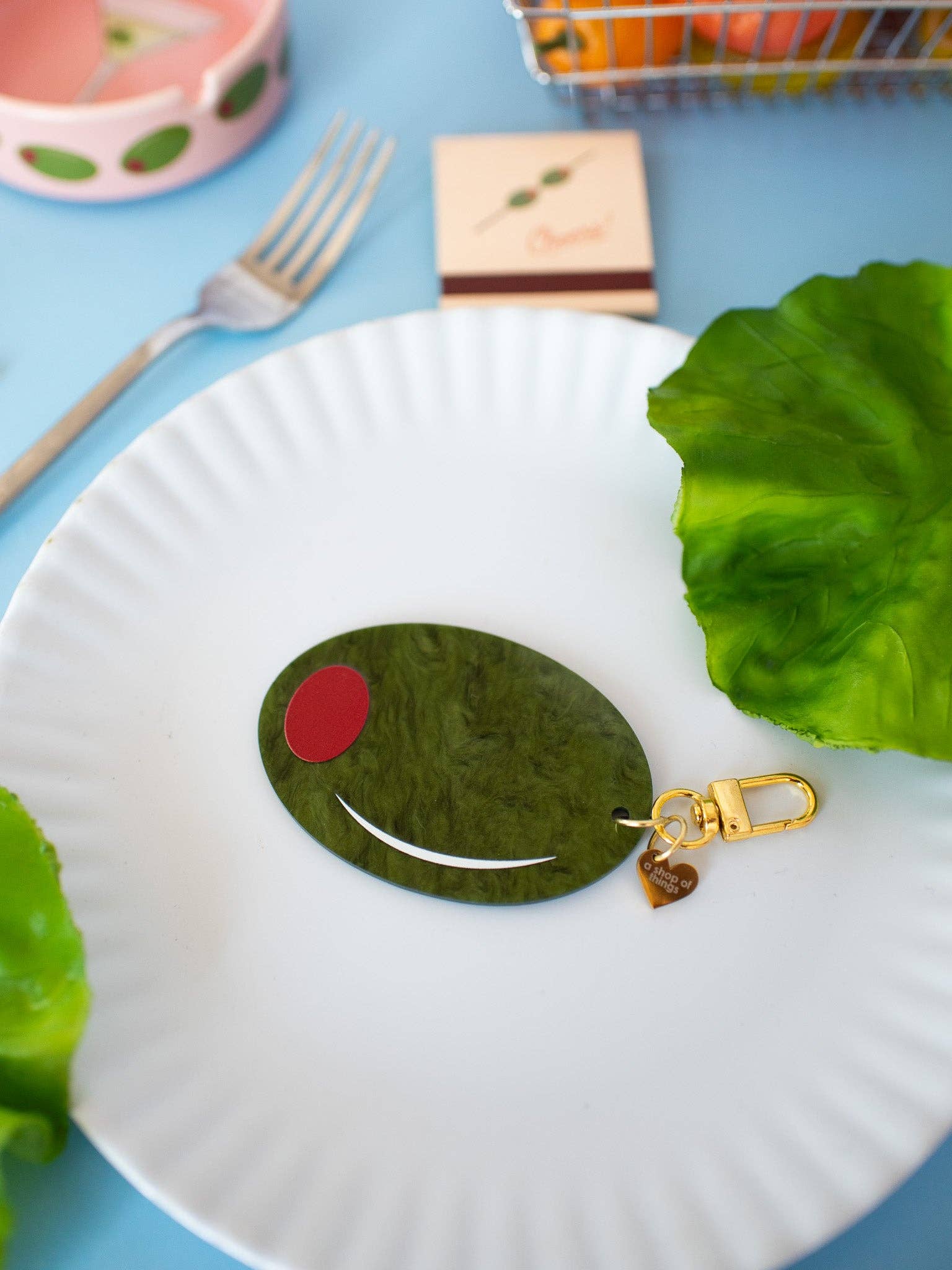 Green pimento olive shaped keychain with gold hardware and a heart-shaped gold charm that reads "a shop of things" 