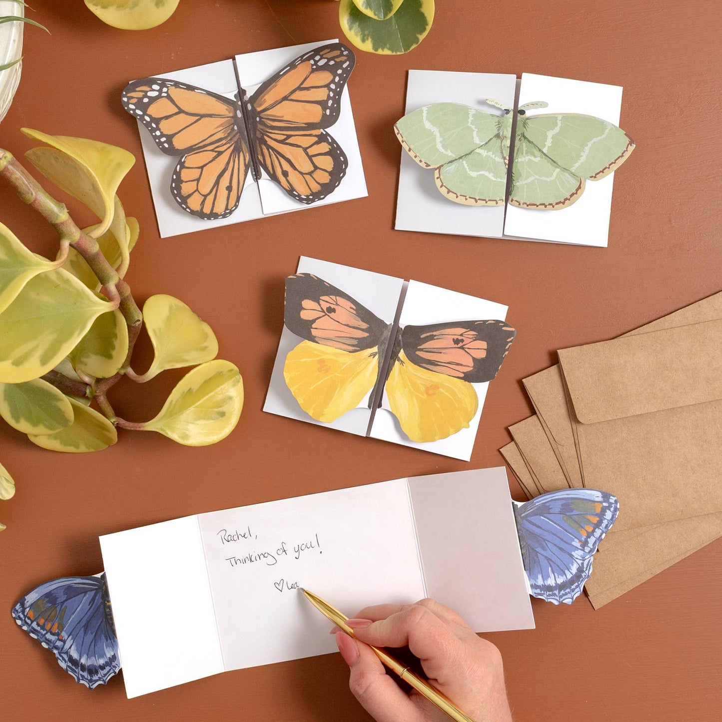 Butterfly-themed greeting cards and envelopes on a brown surface with a hand writing a card.