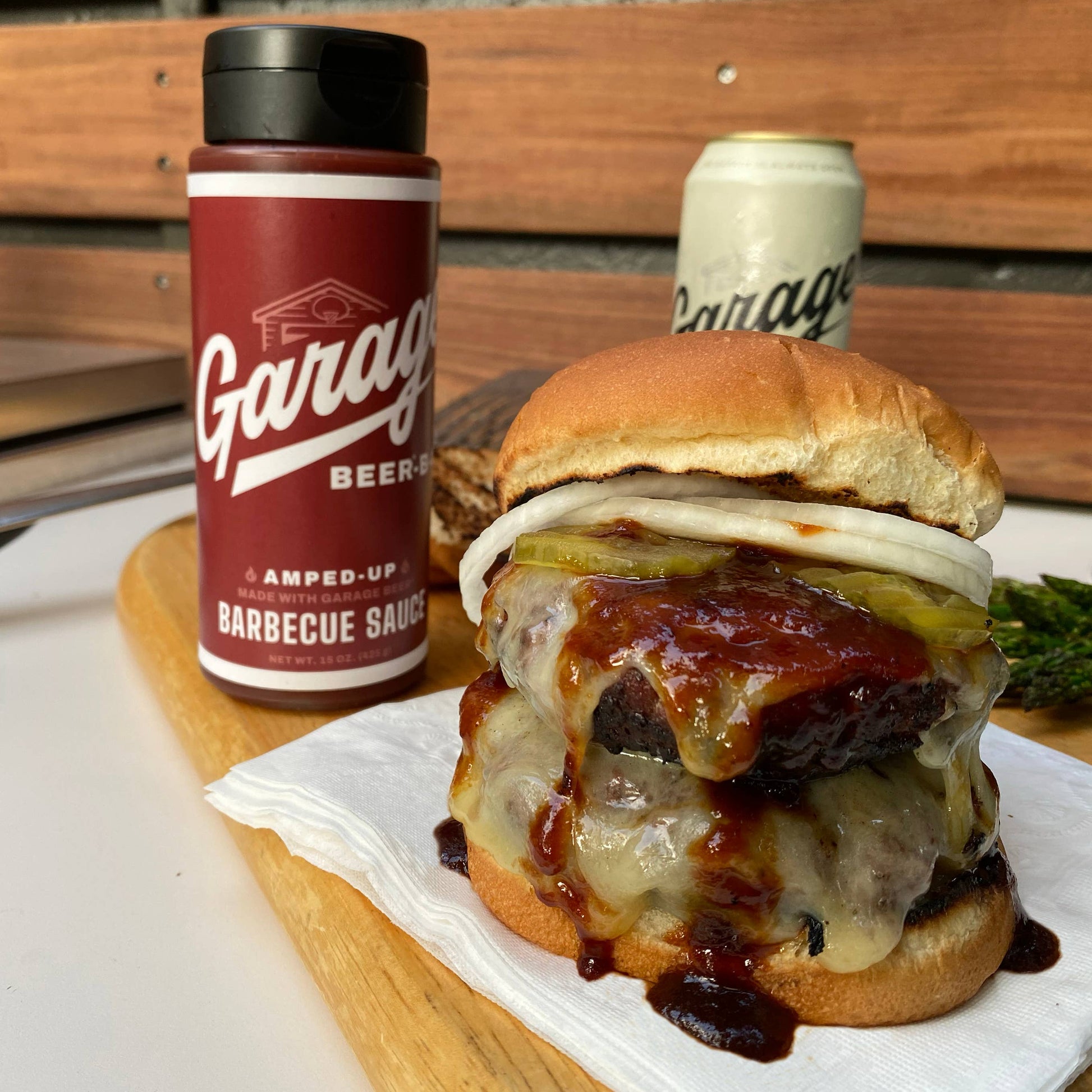 Hamburger with toppings on a wooden board next to a bottle of Garage Amped-Up Barbecue Sauce and a can of Garage beer.