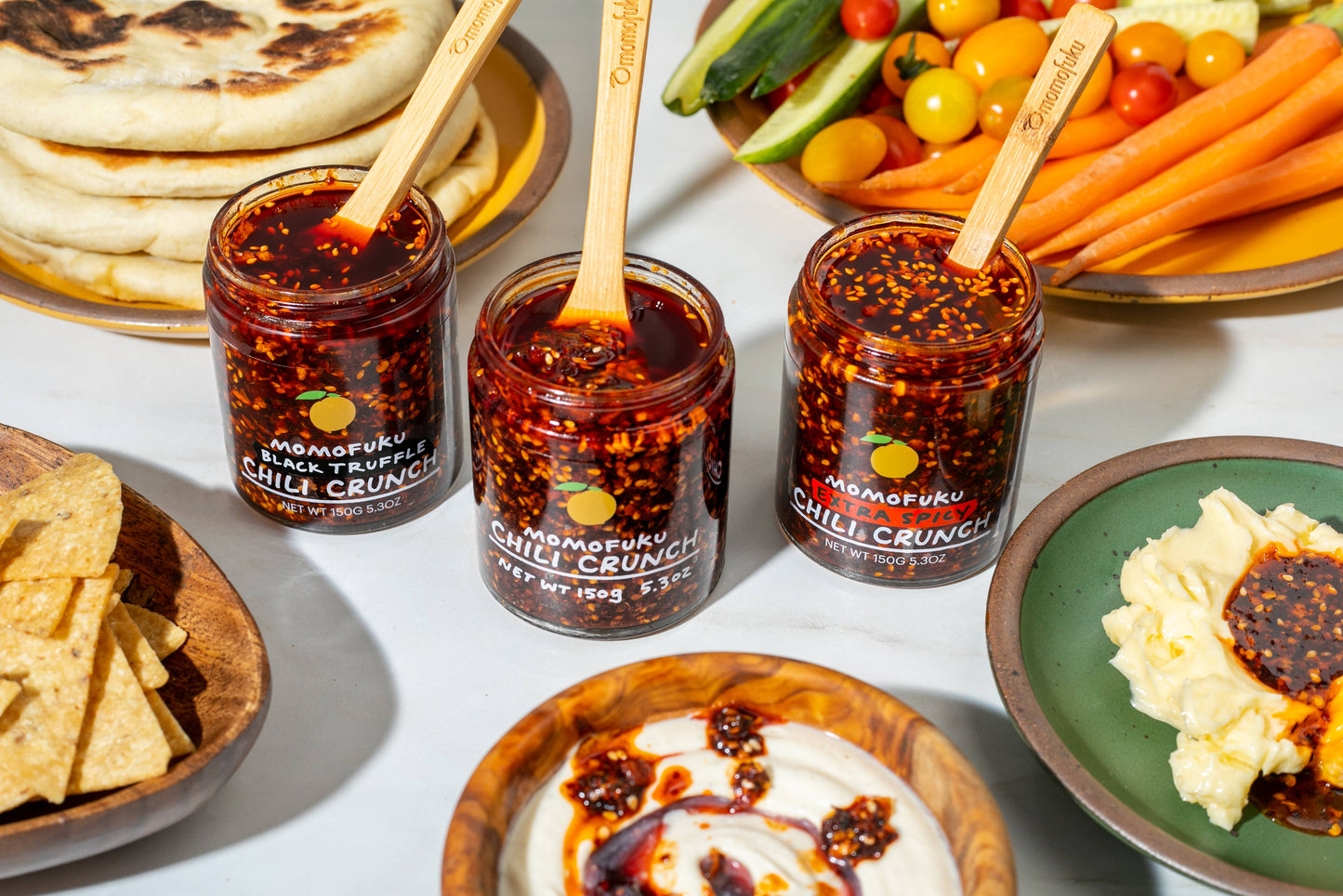Three jars of Momofuku Chilli Crunch with spoons, surrounded by snacks and vegetables on a table.