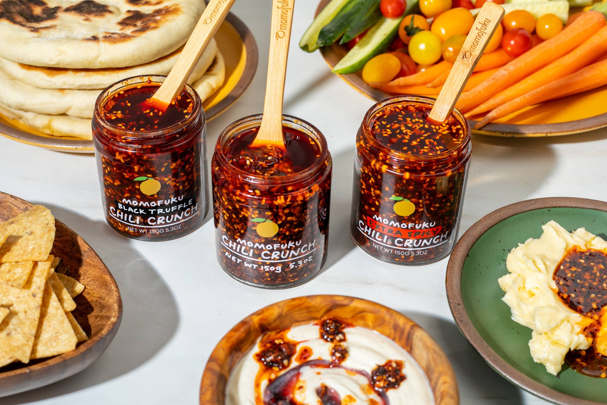 Three jars of Momofuku Chilli Crunch with spoons, surrounded by snacks and vegetables on a table.