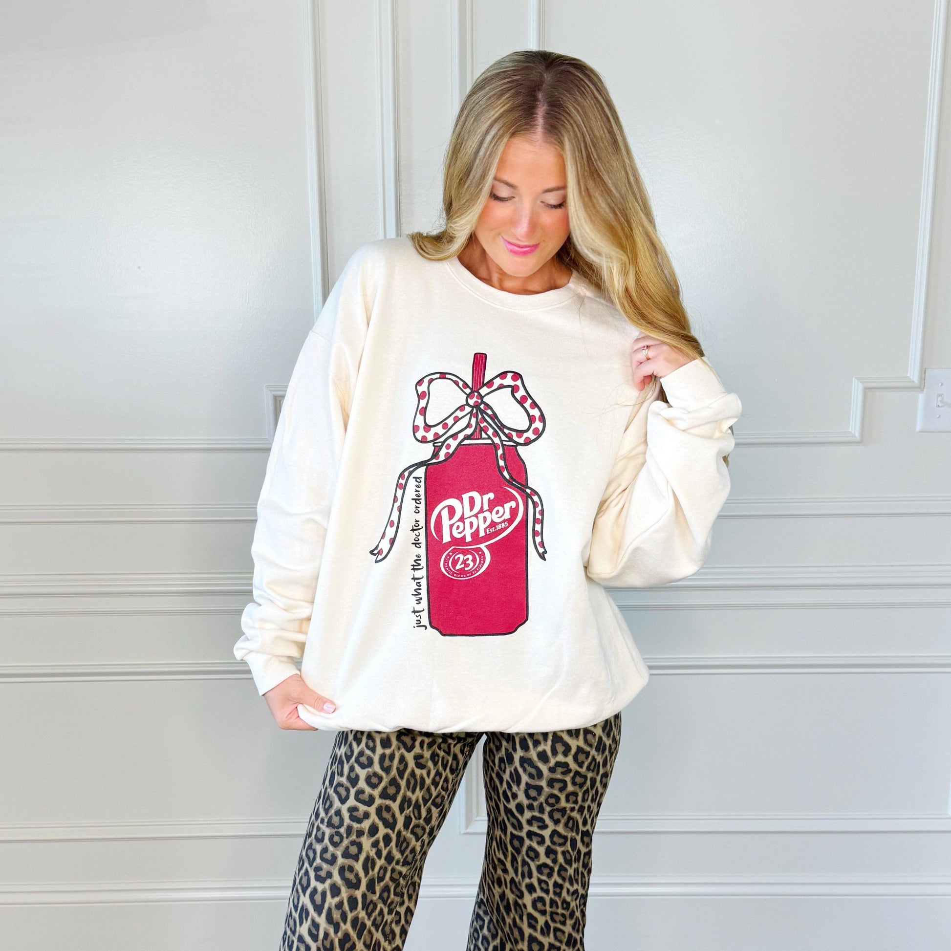 Woman wearing an off-white sweatshirt with a Dr Pepper can with polka-dot bow design,  reading just what the doctor ordered" standing against a white wall.