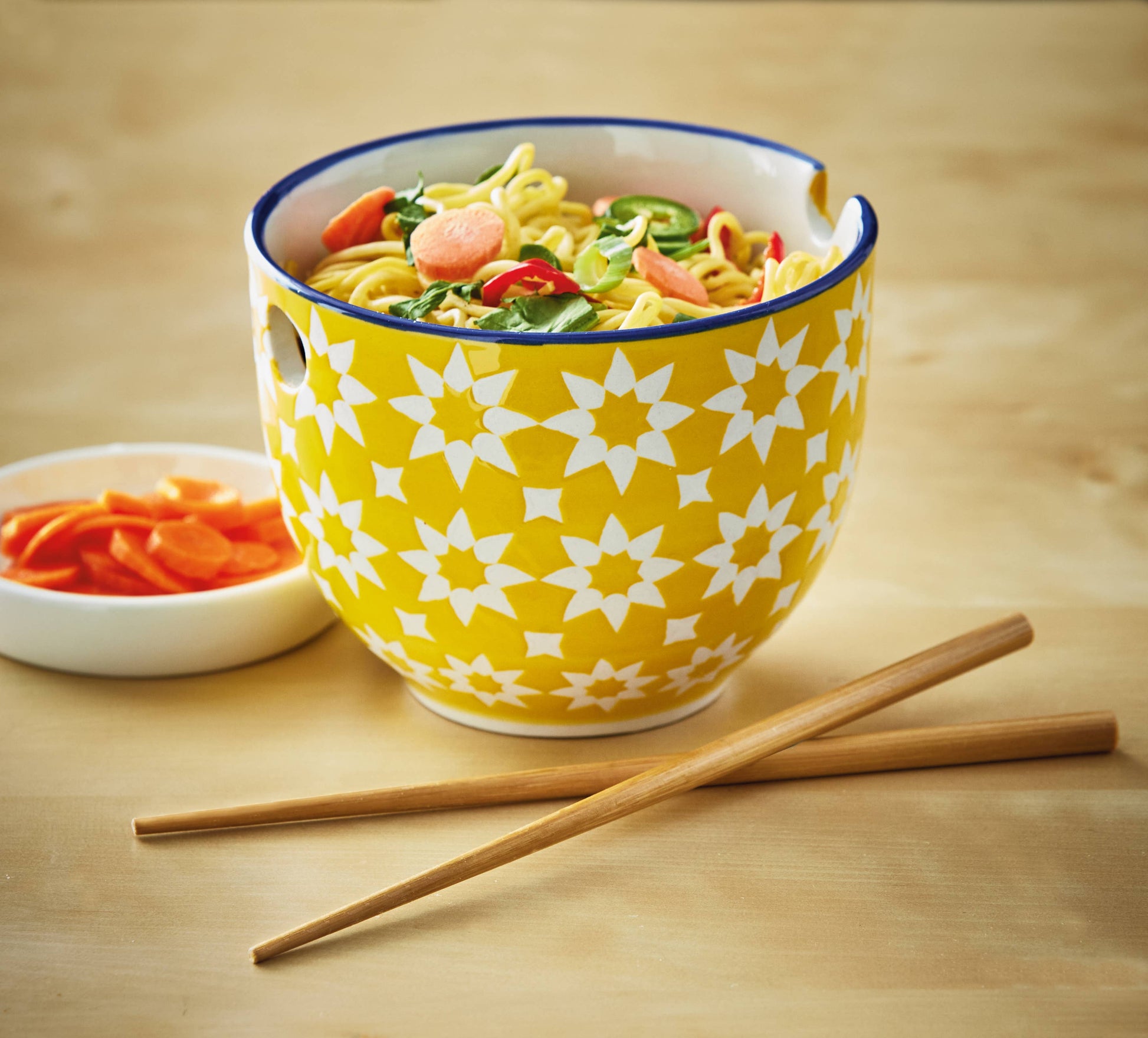 A bowl of ramen with chopsticks and a dish of carrotson a subtle wooden tabletop. Noodle bowl is yellow with white quilted flowers and a blue edge.