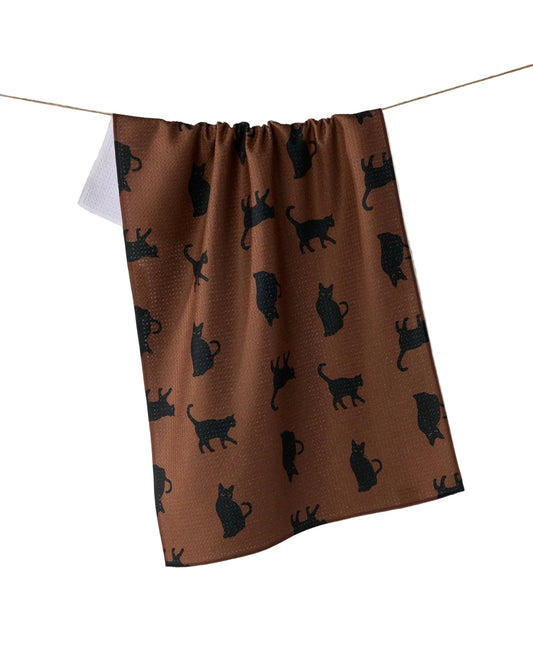 A brown kitchen towel with a black cat silhouette pattern, hanging from a string against a light gray background.