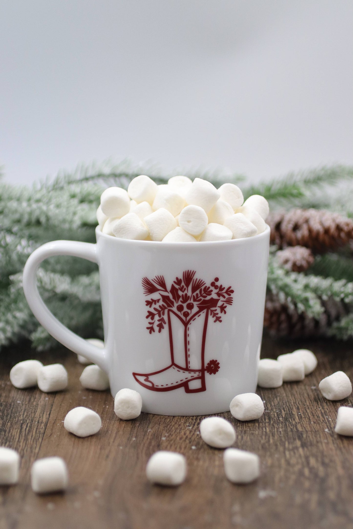 White mug with red tree design filled with marshmallows on a wooden surface with pine cones and branches in the background.
