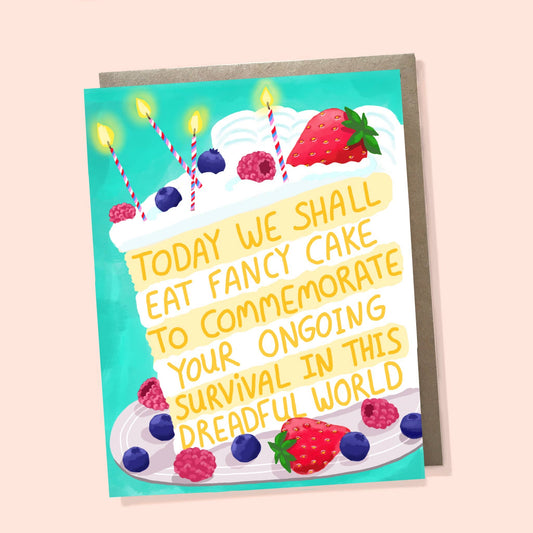 A greeting card featuring a colorful illustration of a birthday cake topped with strawberries, blueberries, and candles, with the text 'TODAY WE SHALL EAT FANCY CAKE TO COMMEMORATE YOUR ONGOING SURVIVAL IN THIS DREADFUL WORLD'. The card is shown with a brown envelope against a pale peach background.