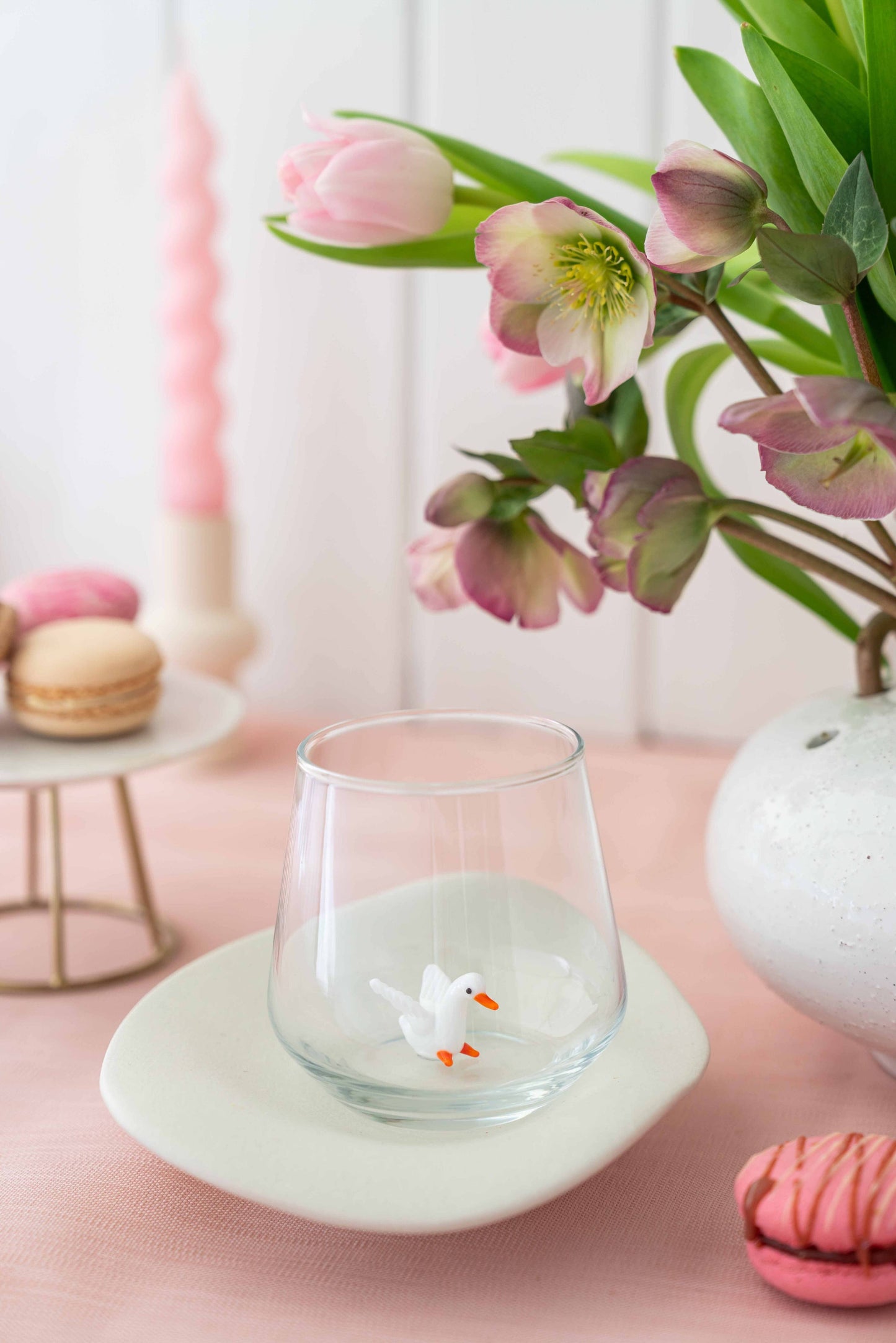 Clear glass with a small figurine on a pink surface with flowers and macarons in the background