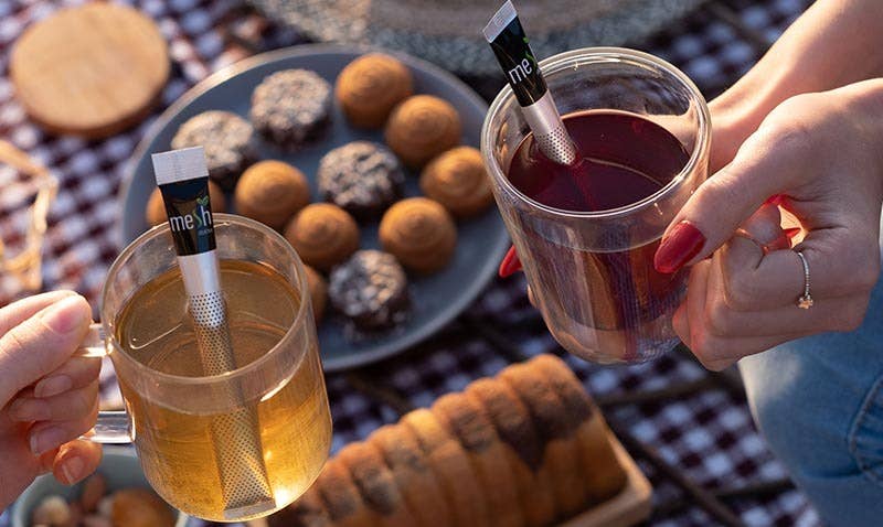 Two people holding cups of tea with Numi tea bags, surrounded by pastries on a checkered tablecloth.