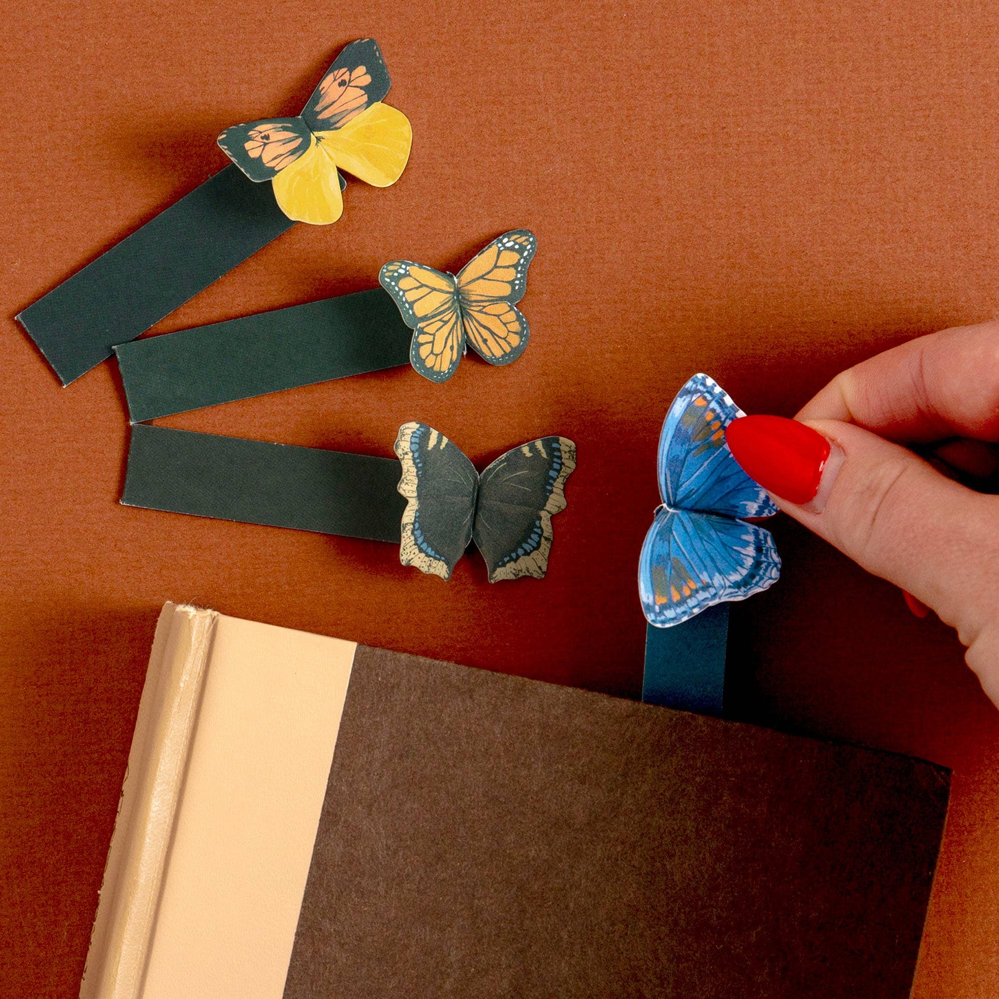 Butterfly-shaped bookmarks on a brown surface with a hand holding a blue bookmark.
