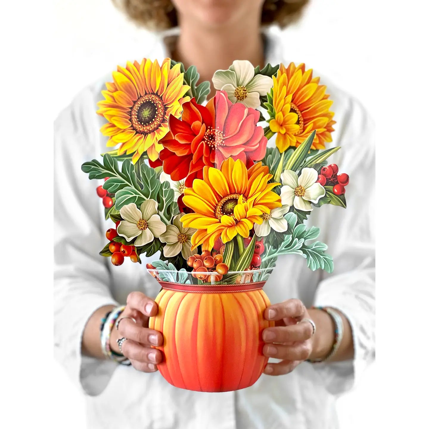 A person in a white shirt holds a decorative floral arrangement in a pumpkin-shaped vase. The bouquet features large yellow sunflowers, orange and red flowers, white blossoms, green leaves, and small red berries.