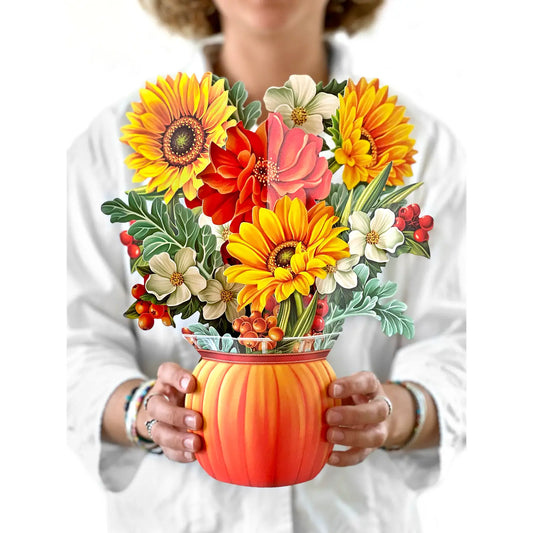 A person in a white shirt holds a decorative floral arrangement in a pumpkin-shaped vase. The bouquet features large yellow sunflowers, orange and red flowers, white blossoms, green leaves, and small red berries.