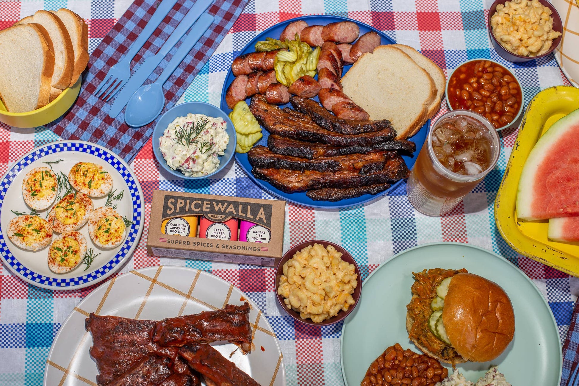 Outdoor picnic setup with plates of food, drinks, and Spicewalla seasoning on a checkered tablecloth.