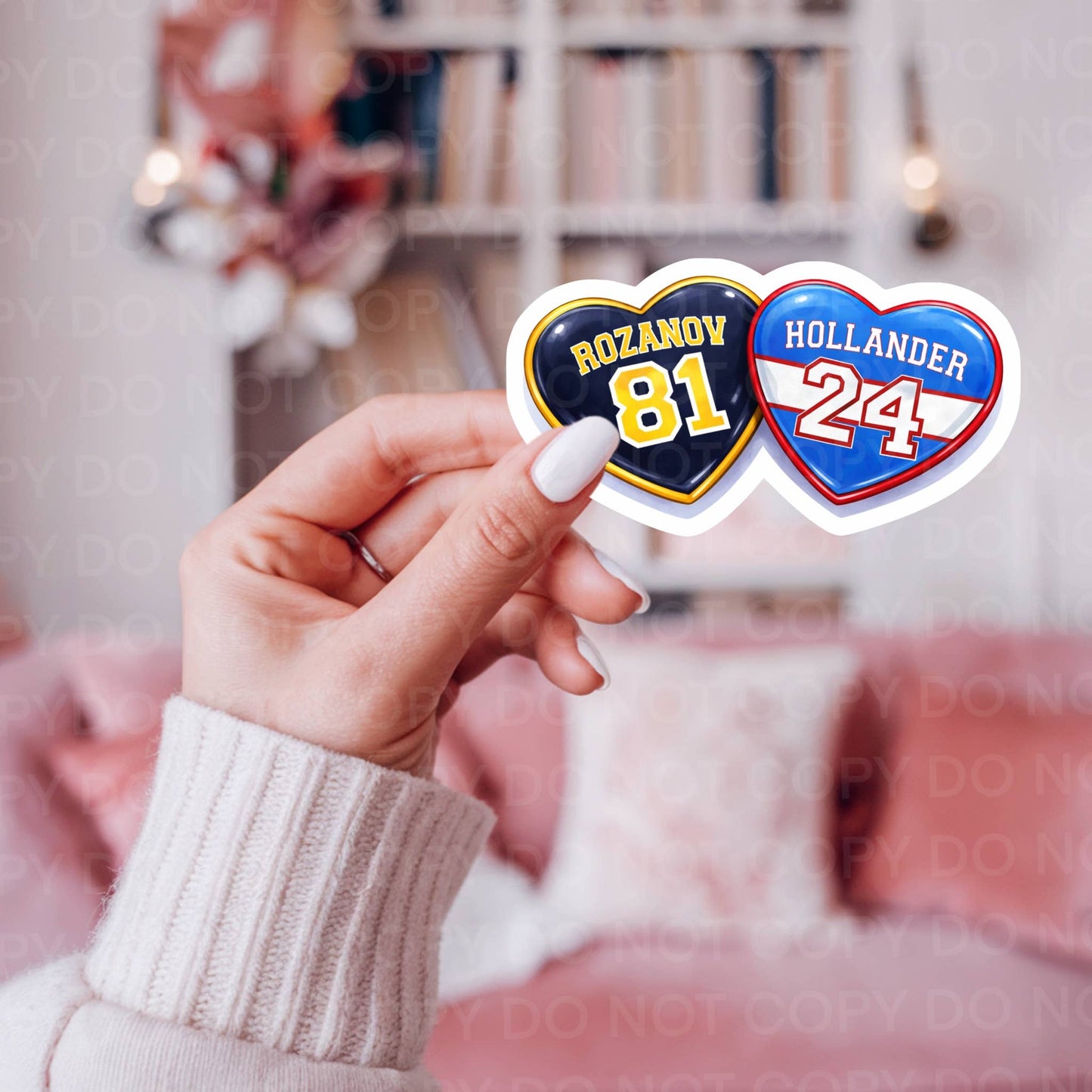 Two heart-shaped stickers with names and numbers held by a hand in a blurred indoor setting.