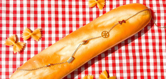 A gold necklace with pasta shaped charms placed on a loaf of bread atop a white and red checkered tablecloth