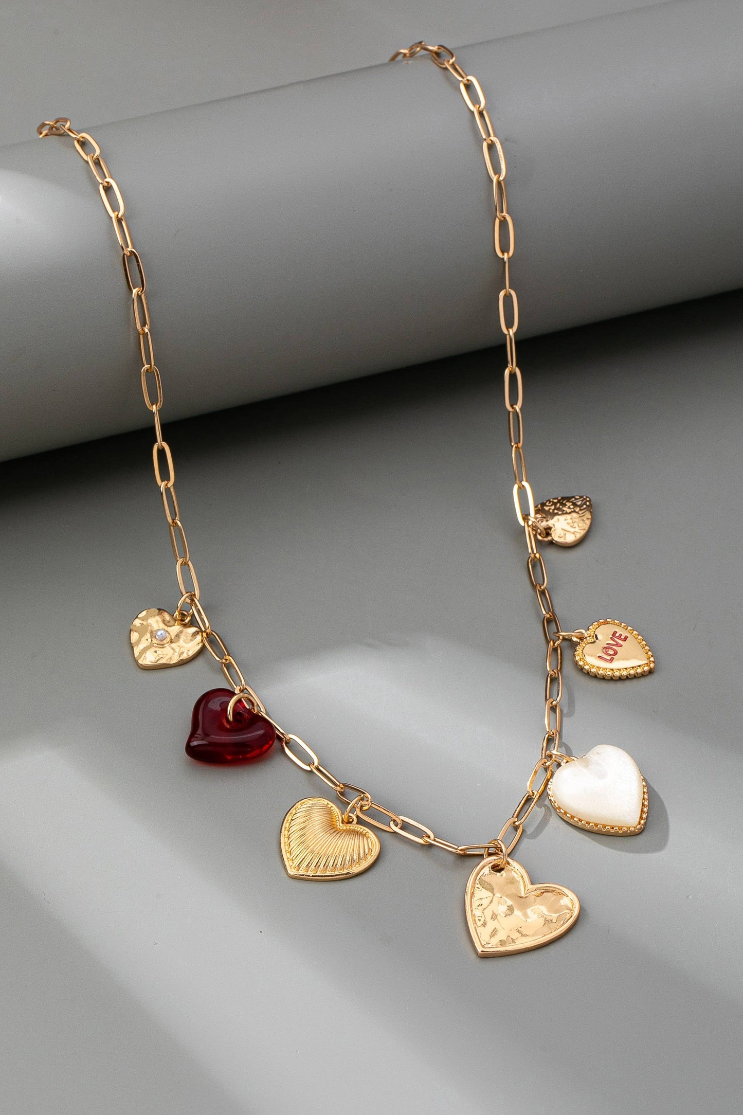 Gold chain link necklace with 5 different heart shaped gold charms, 1 pearl heart charm, and one clear red charm, on a grey background