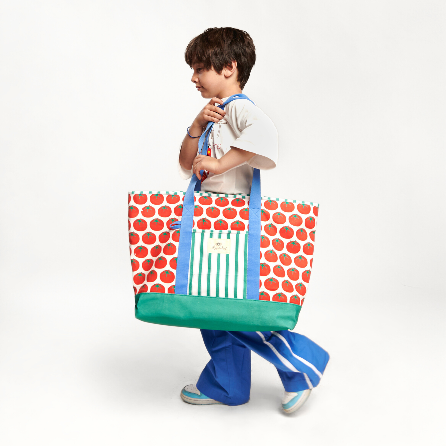 Child holding a colorful tote bag with tomatoes pattern, blue handles and green base on a white background