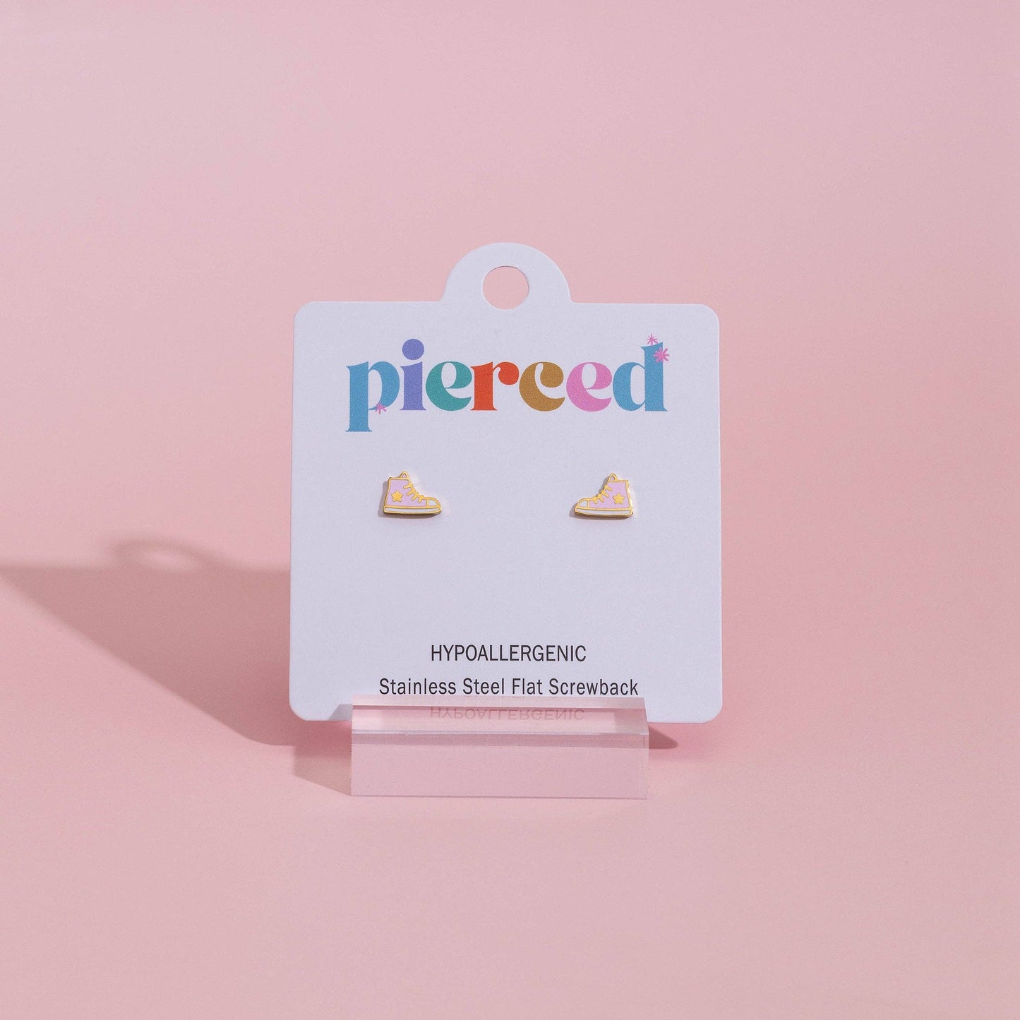 A pair of small flat stud earrings displayed on a white jewelry card labeled 'pierced', standing on a clear acrylic easel against a soft pink background.