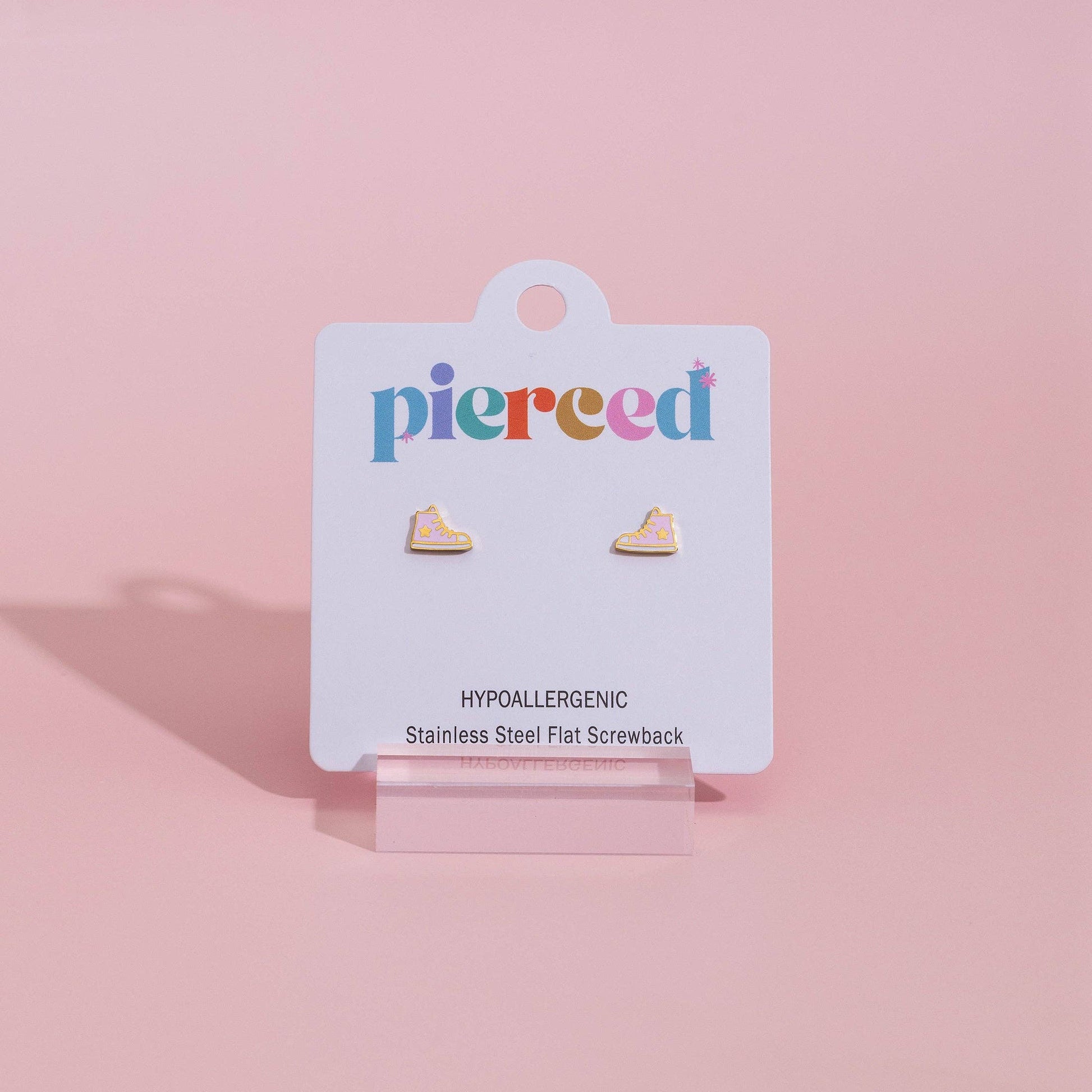 A pair of small flat stud earrings displayed on a white jewelry card labeled 'pierced', standing on a clear acrylic easel against a soft pink background.