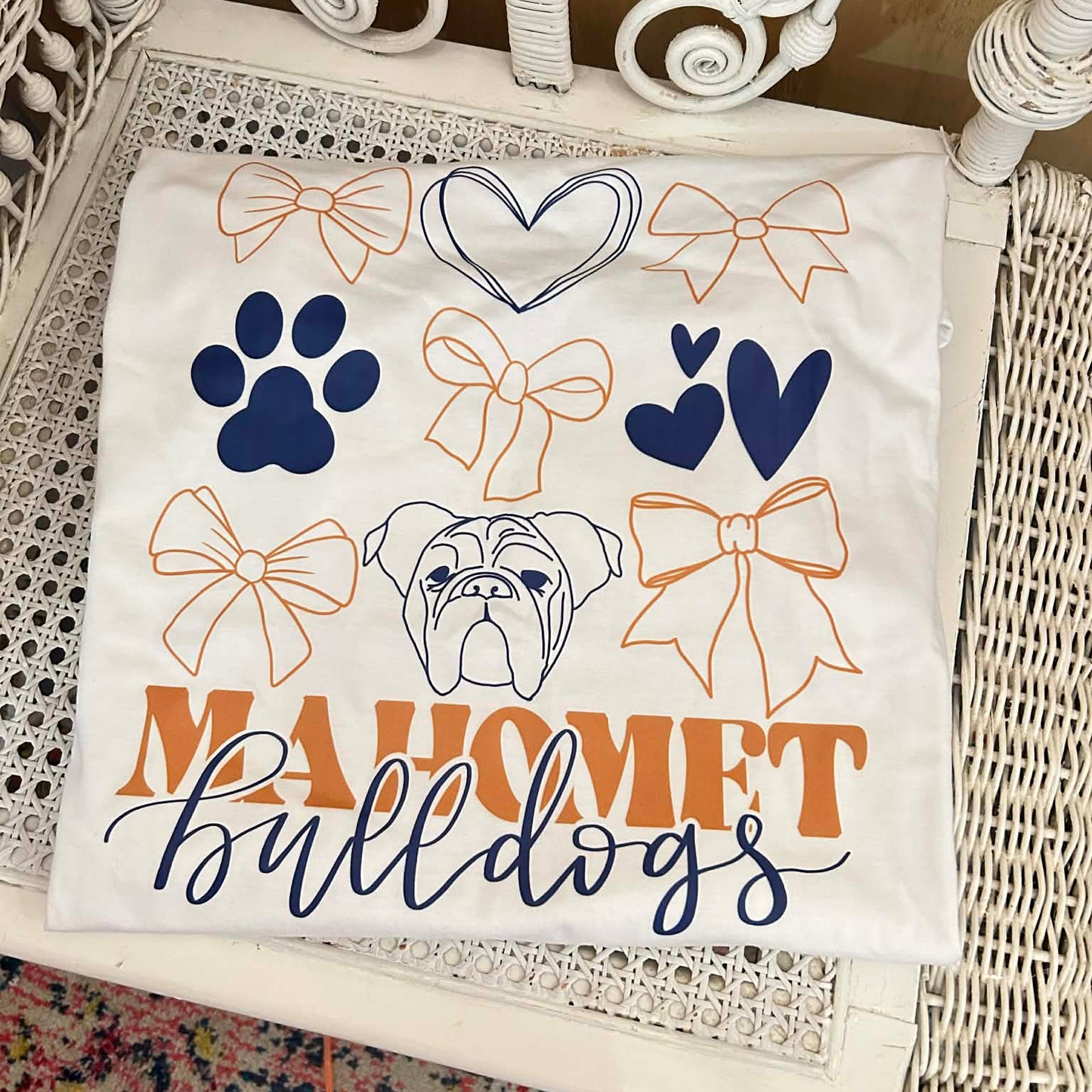 White t-shirt with bows, hearts, paw prints and a bulldog graphic with Mahomet Bulldogs text sitting folded on a chair