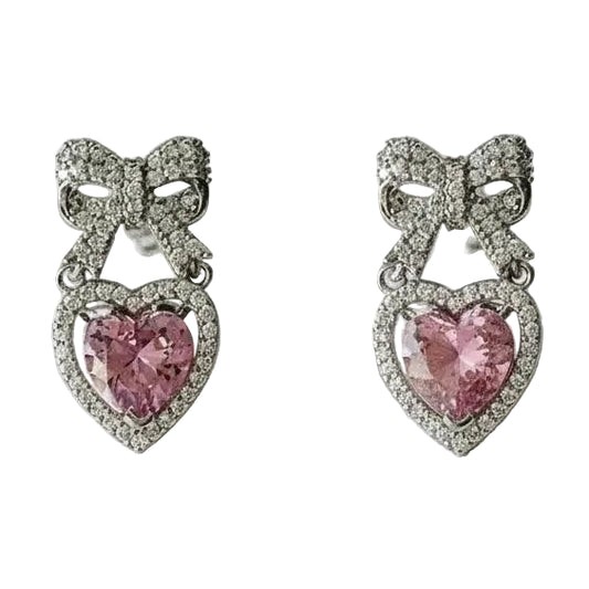 Pair of earrings with pink heart-shaped gemstone in the center, with a rhinestone encrusted bow design above on a white background