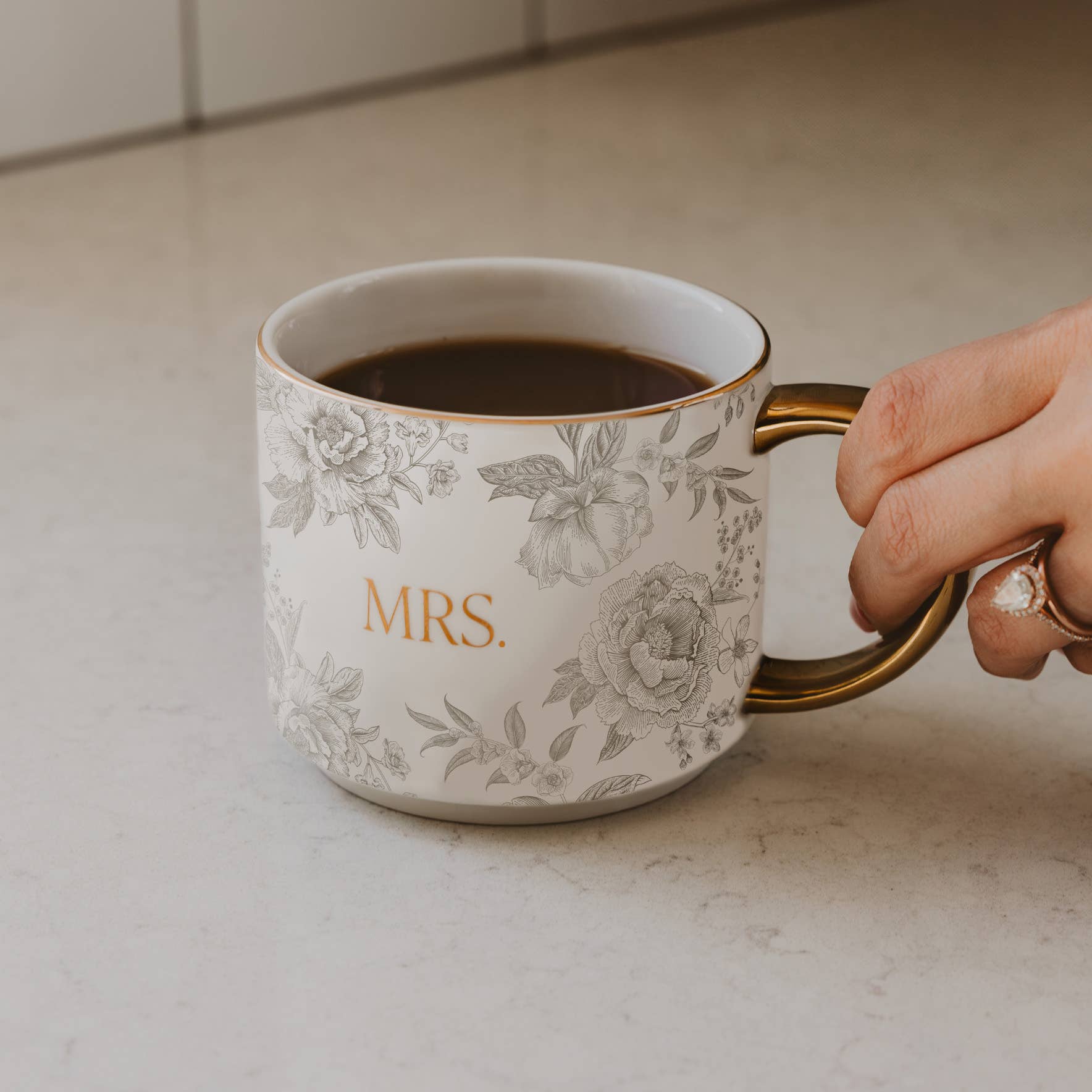 Person holding a floral-patterned mug with 'MRS' on a countertop