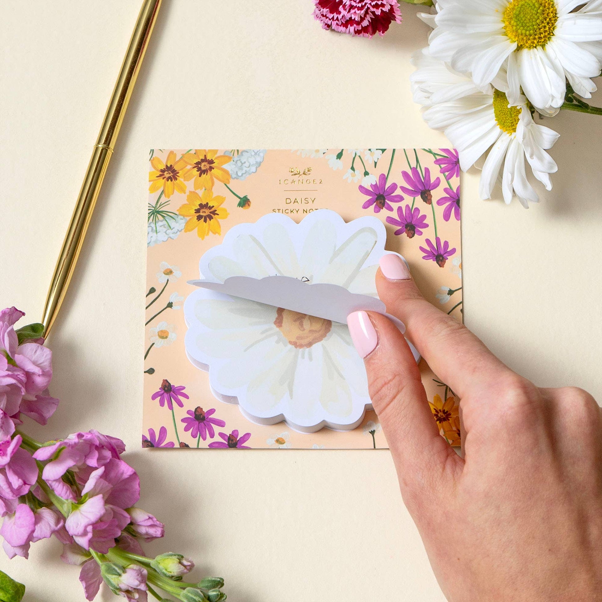 Hand holding a white flower-shaped sticky note over a floral-themed packaging with flowers around.