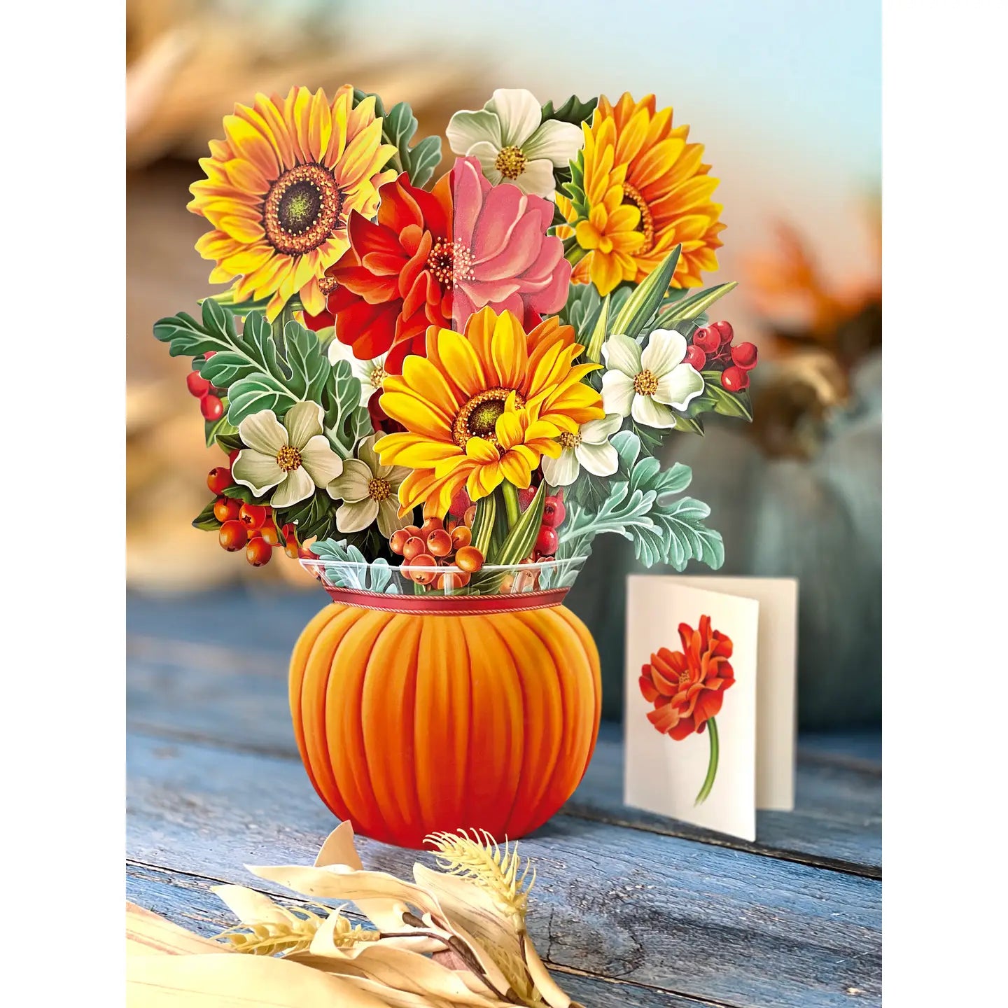 A decorative floral arrangement in a ribbed orange pumpkin-shaped vase, featuring yellow sunflowers, red and pink flowers, white blossoms, green leaves, and small red berries, displayed on a rustic blue wooden surface with a greeting card and dried corn husks nearby.
