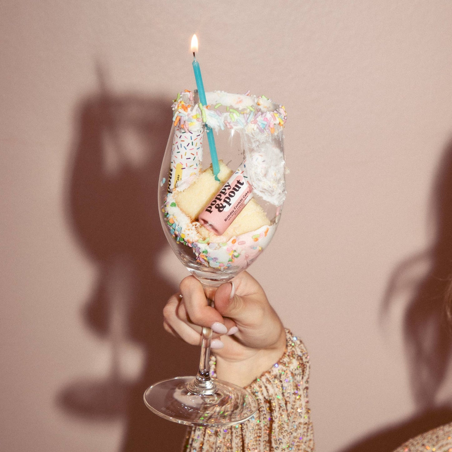 Wine glass filled with cake, candles, and sprinkles held by a person against a plain background