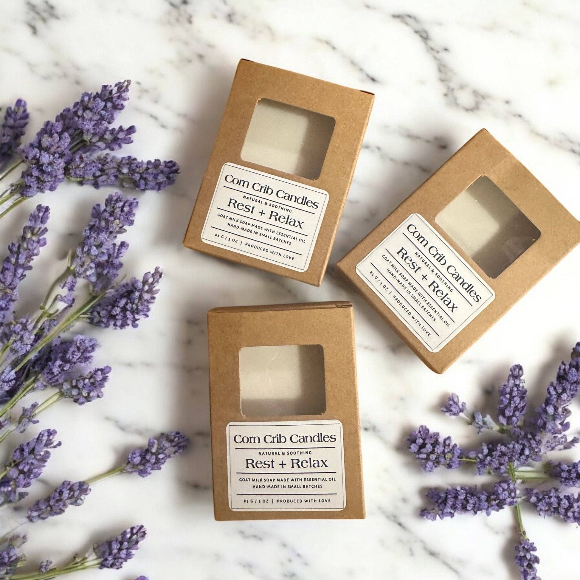Three boxes of Corn Crib Candles labeled 'Rest + Relax' on a marble surface with lavender flowers.