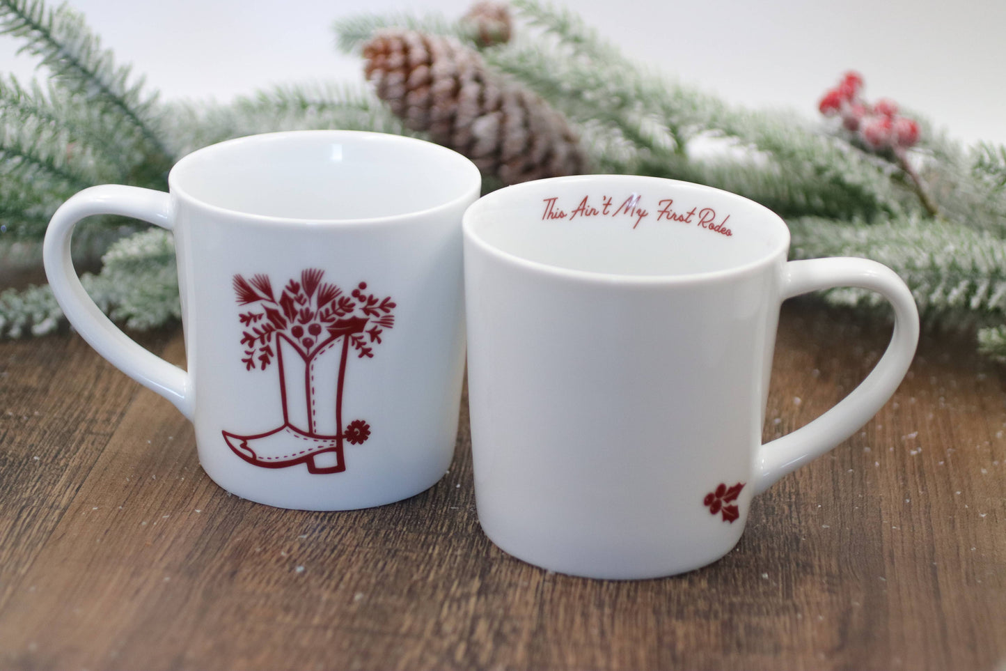 Two white mugs with red designs on a wooden surface with a Christmas-themed background.