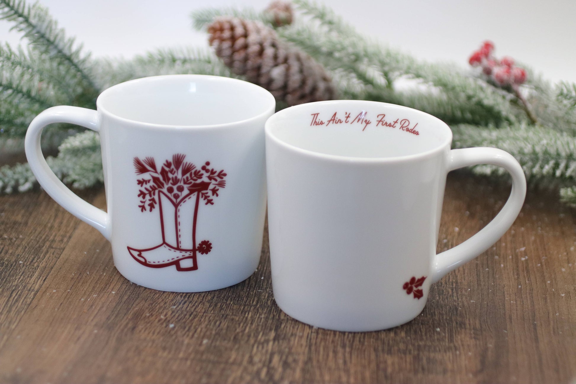 Two white mugs with red designs on a wooden surface with a Christmas-themed background.