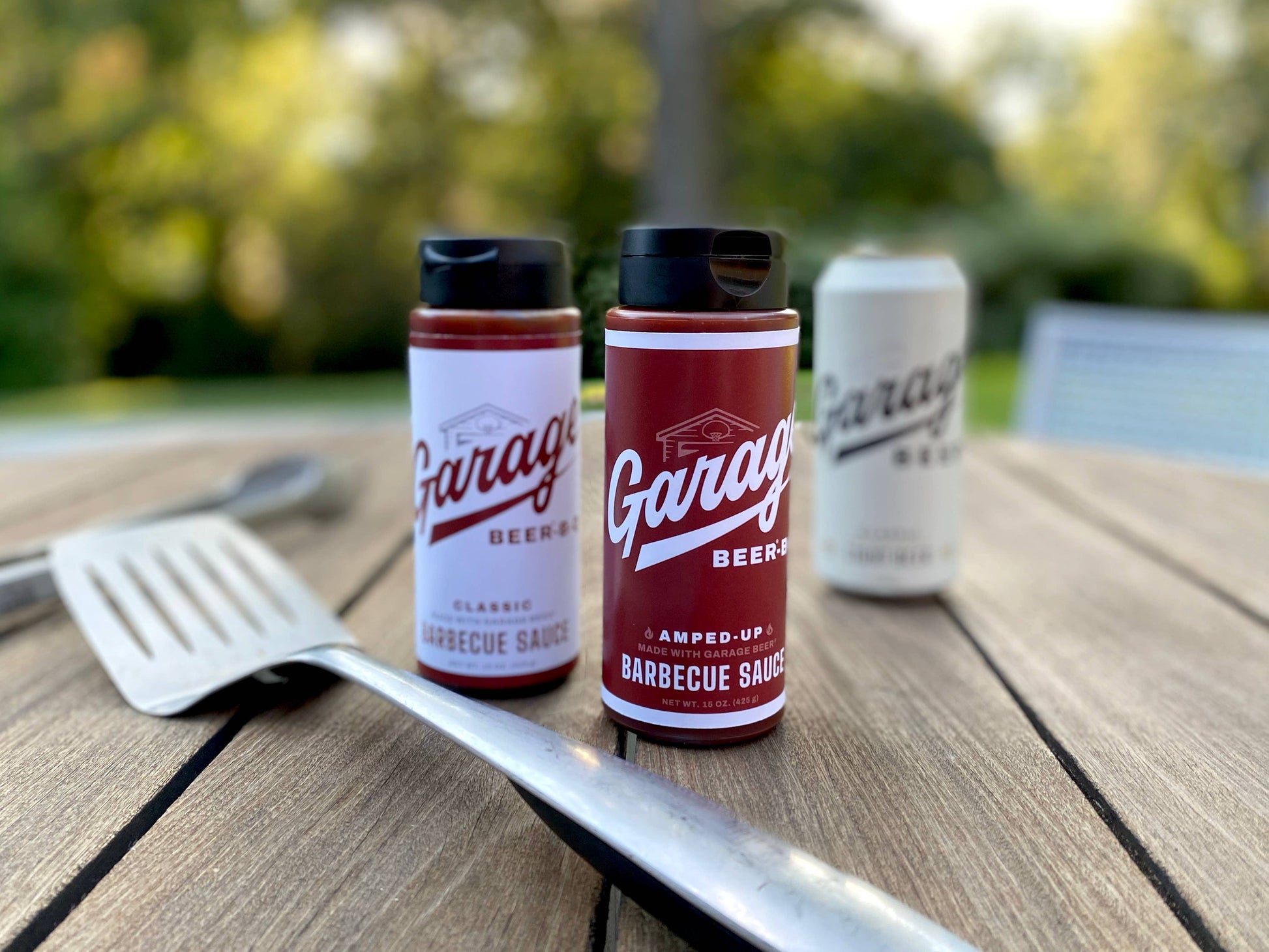 Two bottles of Garage Beer Company barbecue sauce on a wooden table with a blurred outdoor background.