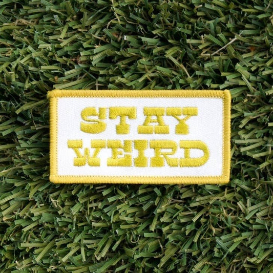 A rectangular embroidered patch with the words 'STAY WEIRD' in yellow lettering on a white background, placed on green grass.