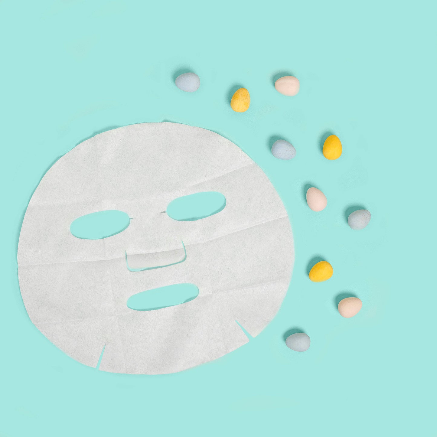 White face mask sheet on a light blue background with colorful easter eggs around it