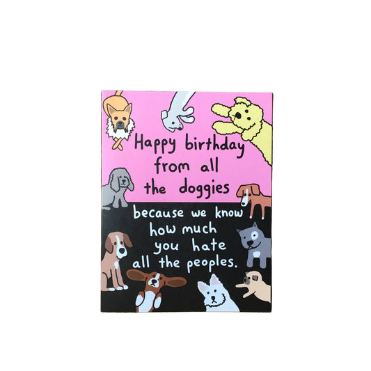 A square greeting card with a pink top half and black bottom half, featuring cartoon dog illustrations and the text 'Happy birthday from all the doggies because we know how much you hate all the peoples.' The card is propped against a rustic brick wall on a wooden surface.