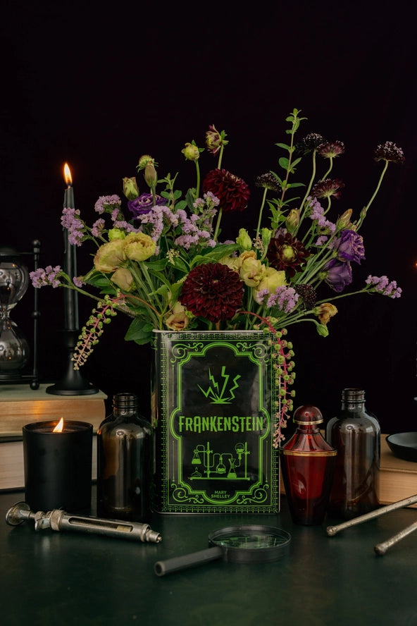 Decorative setup with 'Frankenstein' book, flowers, candles, and bottles on a dark background