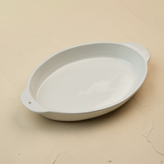 A white oval ceramic baking dish with side handles, photographed on a light beige textured surface.